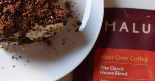 A square slice of tiramisu made with Malu's specialty pour over coffee is served on a plate beside a packet of Malu specialty coffee 